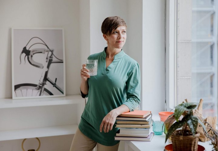 Woman standing by a window holding a glass of water, representing healthy lifestyle factors that may contribute to early menopause.